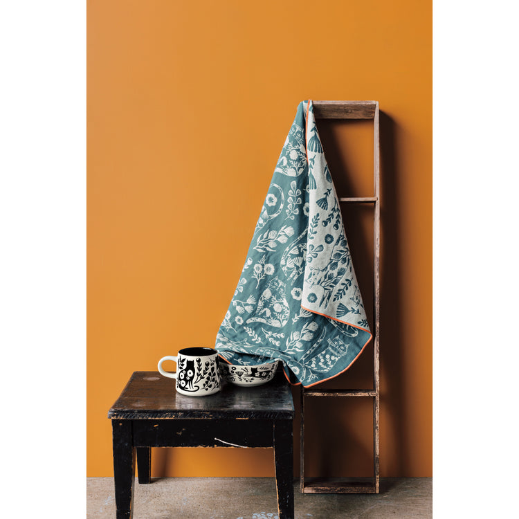 A Danica Studio Double Cloth Catbloom dishtowel with cats and flowers on it next to an Imprint bowl and mug in a matching design, in front of an orange wall.