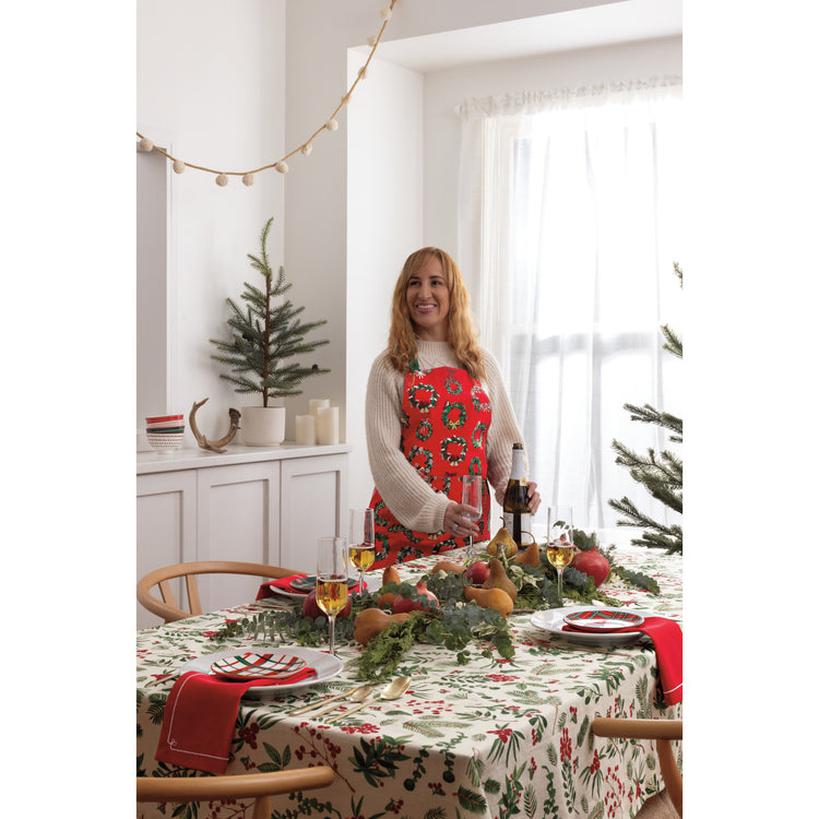 A woman in a red Now Designs Wreaths apron standing in front of a Christmas table with a Winterberry tablecloth.