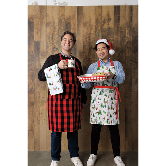 Two men in Now Designs Christmas aprons posing for a picture. One is wearing a black and red Buffalo Check Mighty Apron and holding a printed dishtowel and a mug, and one is wearing a Santa's Reindeer apron and holding a baking dish while wearing a santa hat.