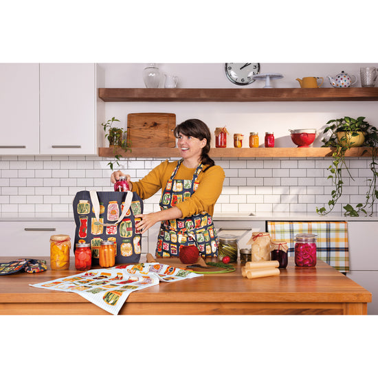 A woman in a Now Designs Pickling apron with jars on it packing food into a Pickling tote bag with jars and cans on the table.