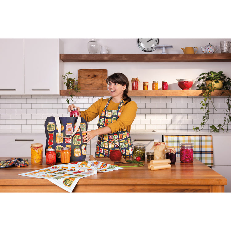 A woman in a Now Designs Pickling apron with jars on it packing food into a Pickling tote bag with jars and cans on the table.