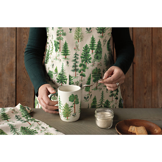 A woman wearing a Now Designs apron with a Woodland tree print, putting sugar into a matching coffee mug with trees on it.
