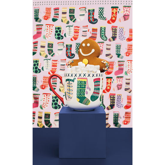 A Danica Jubilee Winter Woolens mug and dishtowel set with a gingerbread man sitting in the mug. The designs on the printed dishtowel and mug feature Christmas stocking and socks.