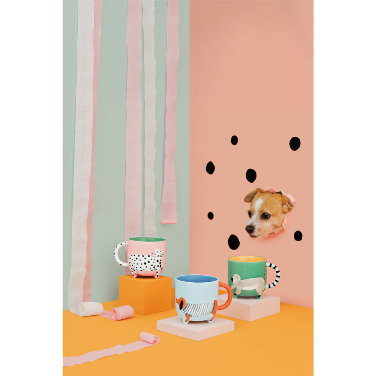 Critter mugs by Danica Jubilee with polka dots and stripes featuring a dog, a cat, and a monkey. There is a dog popping out of the pink wall in the background.