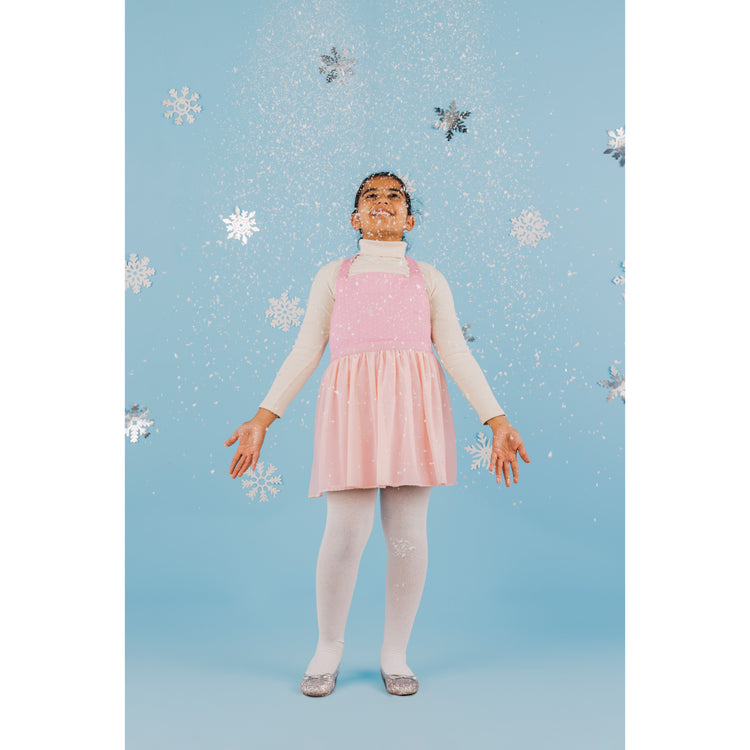 A girl in a Ballerina Kid's Daydream Apron by Danica Jubilee that looks like a pink tutu. She is standing in front of a blue background with snowflakes on it.