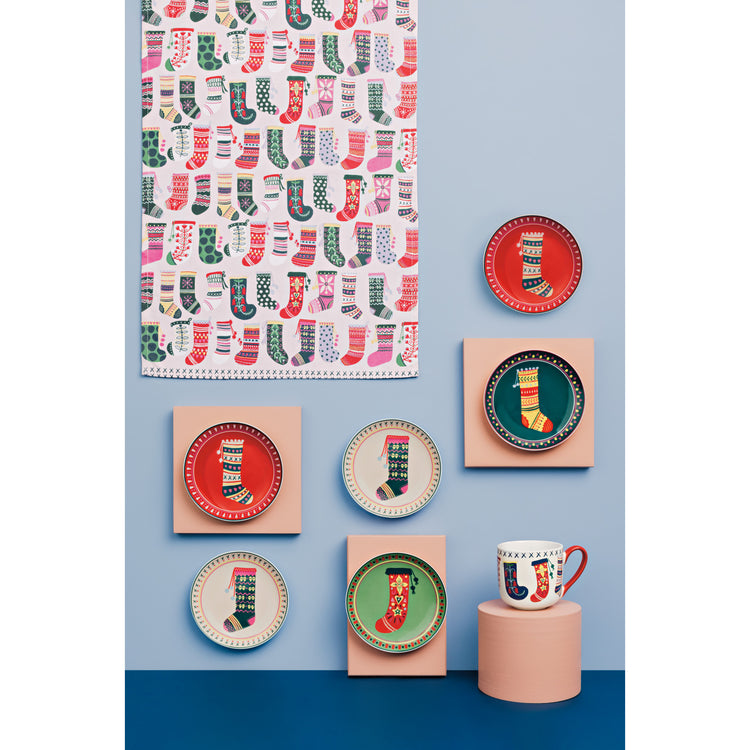 A collection of Winter Woolens appetizer plates, a mug, and a printed dishtowel by Danica Jubilee. The deisgn features Christmas stockings and socks.