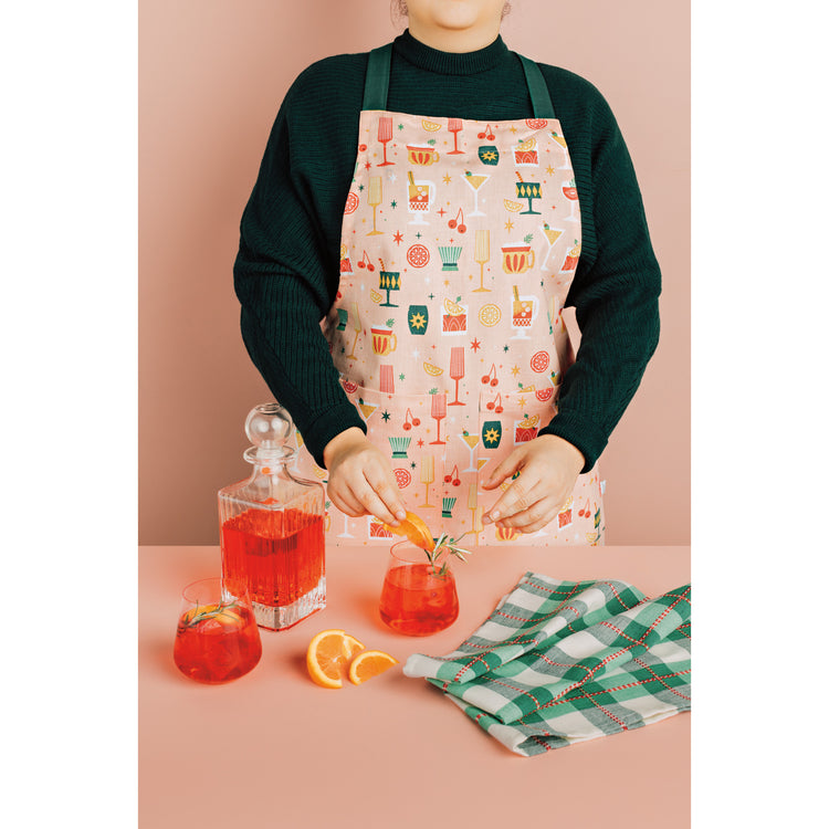 A woman wearing a pink apron from the Danica Jubilee Spirits Bright Christmas Collection is making a cocktail drink.
