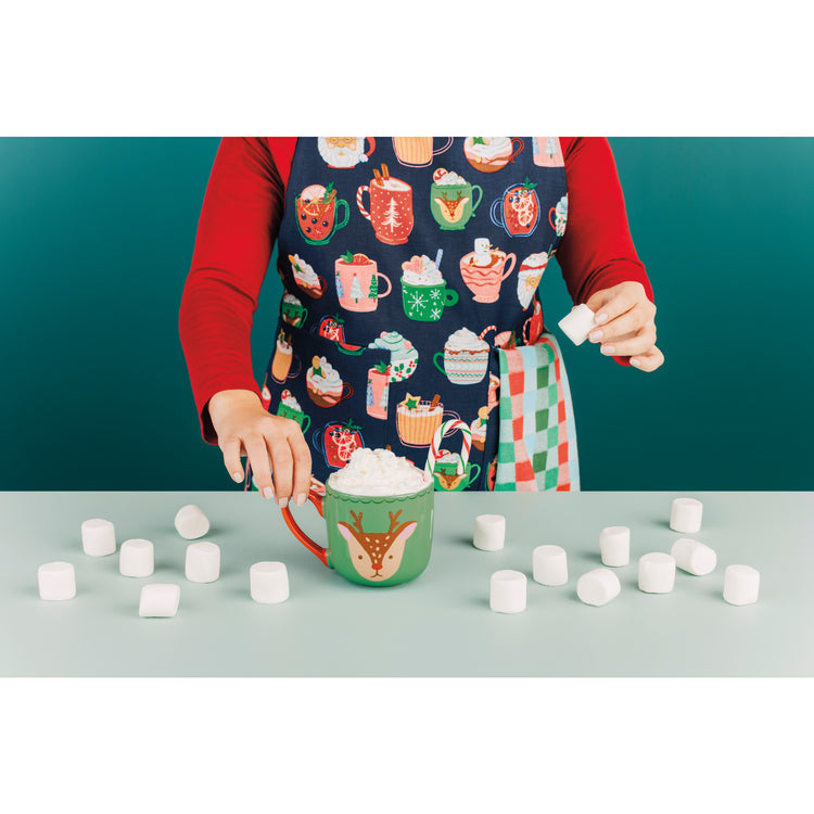 A is woman wearing a printed apron featuring hot drinks from the Cozy Cups Christmas collection by Danica Jubilee. She is putting a marshmallow into a matching green mug with a reindeer's face on it.