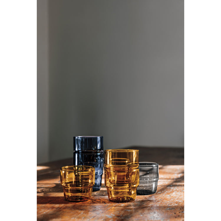 Multicolored Stacked Tumbler glasses in cobalt blue, smoke gray, and amber yellow sitting on a table.