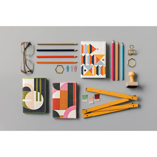 A colorful collection of Danica Studio Formation Printed Notebooks with pens, rulers, and other stationery supplies on a gray surface.