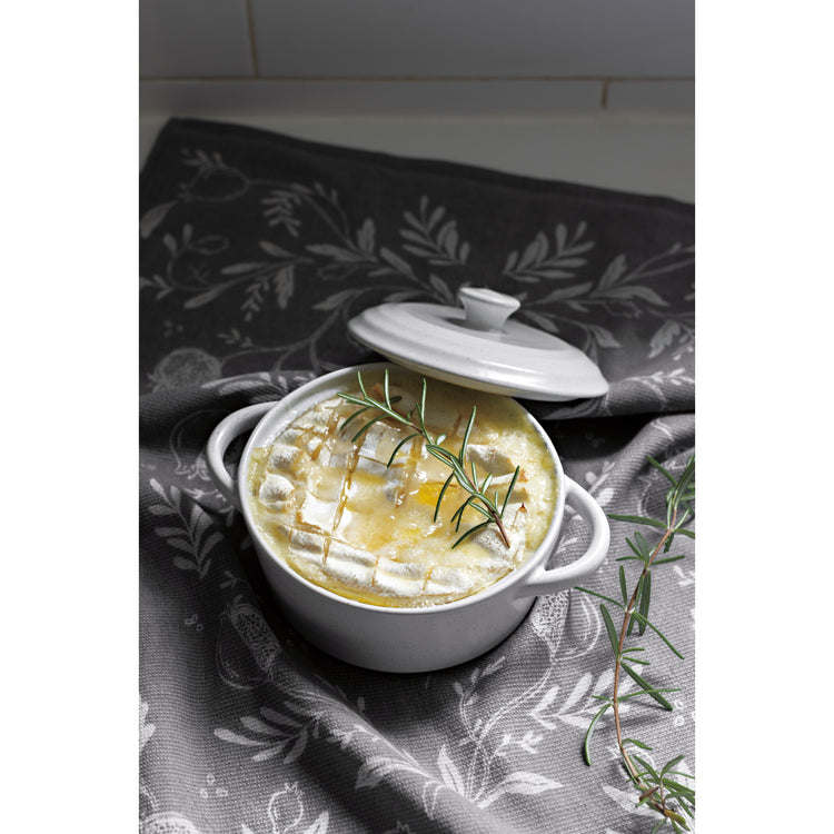 A Now Designs white brie baker dish with a baked brie cheese and a sprig of rosemary on it, sitting on a gray and white printed dishtowel.