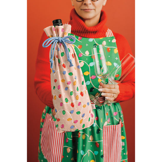 A woman is holding a wine bottle inside a Danica Jubilee wine bag from the Holiday Glow collection and wearing a matching apron with Christmas lights on it.