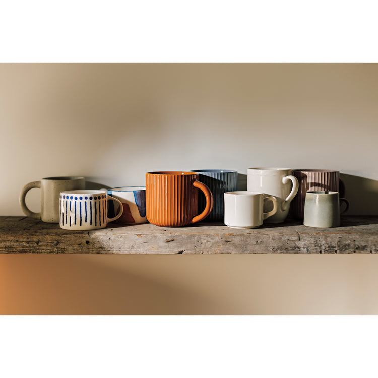 A collection of Heirloom Fluted Mugs in Cognac, Ocean, and Olive Branch sitting on a wooden shelf. There is also an Ivory Provence Mug, a Sprout Mug, a Maison Mug, a Canvas Mug, and a Sage Aquarius Mug.