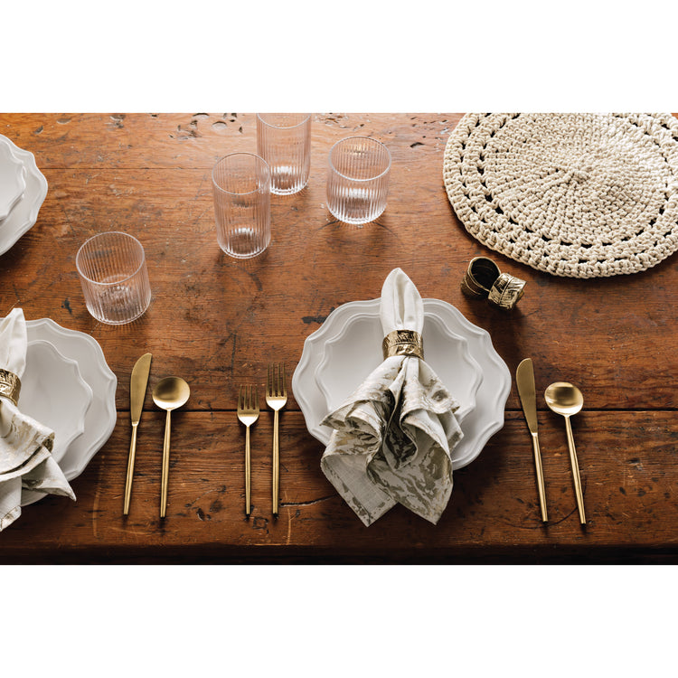 A table setting with Provence dinnerware, matte gold Taper flatware, clear fluted tumbler glasses, Glimmer table linens, a natural knotted placemat, and gold leaf napkin rings on a wooden table.