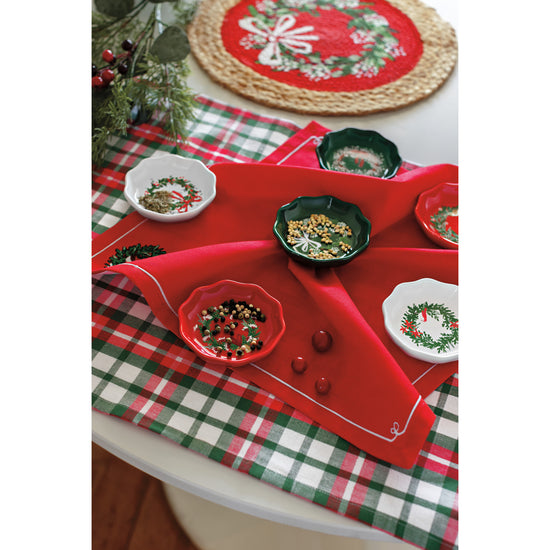 A table with a Now Designs Christmas Wreaths braided placemat, a plaid dishtowel, a red napkin with a wreath on it, and a set of Wreath shaped pinch bowls in red, white, and green.