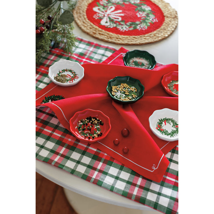 A table with a Now Designs Christmas Wreaths braided placemat, a plaid dishtowel, a red napkin with a wreath on it, and a set of Wreath shaped pinch bowls in red, white, and green.