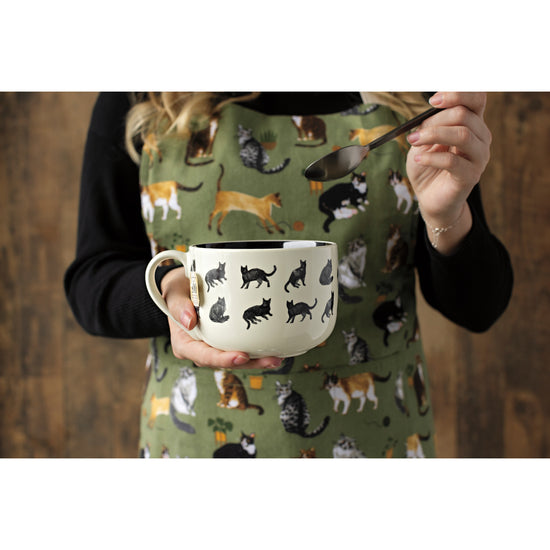 A woman wearing a Now Designs Cat Collective apron holding a cat print latte mug and a spoon.