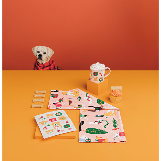 A dog sits next to a table that shows the Danica Jubilee Holiday Hounds Christmas Collection featuring a mug, a printed dishtowel, and a Swedish Sponge Cloth.