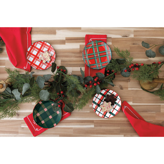 A table with Now Designs assorted plaid appetizer plates, red printed napkins with wreaths on them, and festive Christmas greenery.