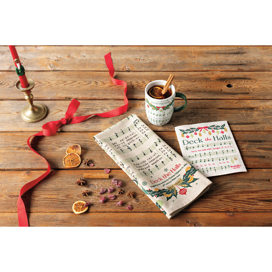 A Now Designs printed dishtowel with sheet music for a Christmas Carol on it and a matching Swedish Sponge Towel and mug next to cinnamon sticks and a candle on a wooden table.