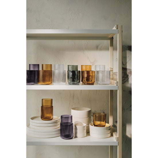 A shelf with a variety of Heirloom Fluted and Bubbled Tumbler Glasses in Smoke gray, Amber yellow, Agave green, and Clear and stacks of ivory Foundation plates and bowls.