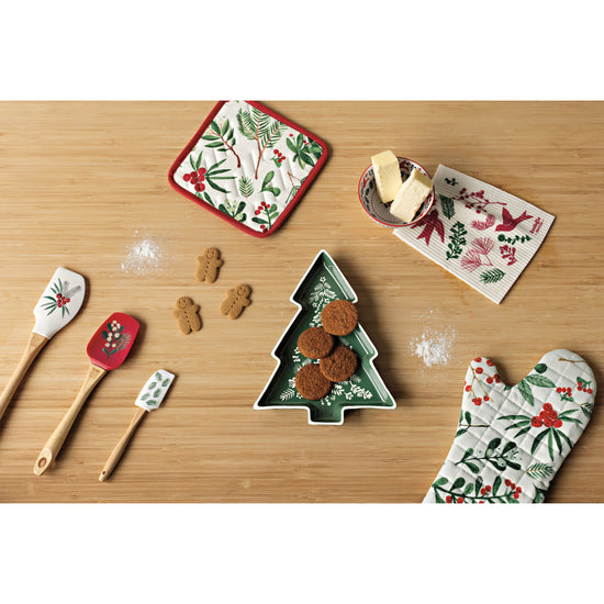 A table with a Now Designs Winterberry shaped tray in the shape of a Christmas tree, an oven mitt, a Swedish Sponge Cloth, a potholder, and a set of spatulas with gingerbread cookies.