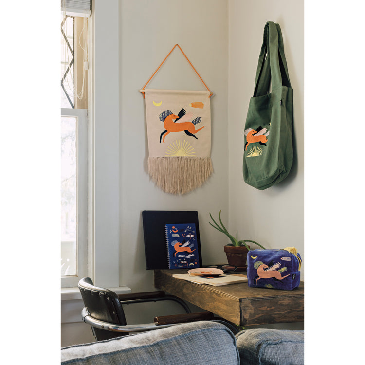 A room with a green Astral tote bag by Danica Studio and matching wall art featuring a pegasus horse hanging on the wall. There is a desk with a blue embroidered pouch and a notebook with the same Astral design.