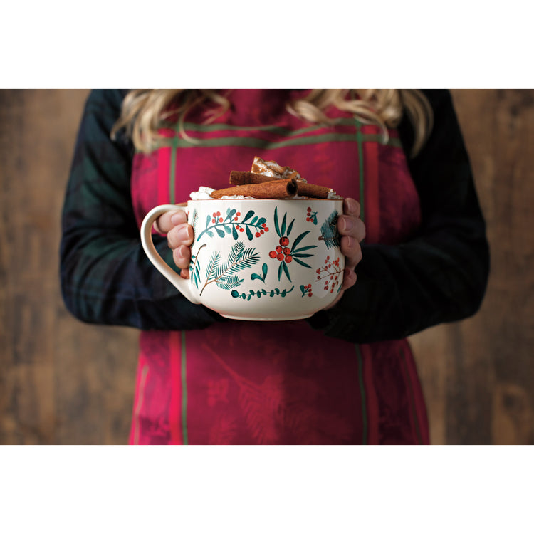 A woman wearing a Now Designs Christmas Winterberry jacquard apron holding a Winterberry print latte mug with hot chocolate and cinnamon.