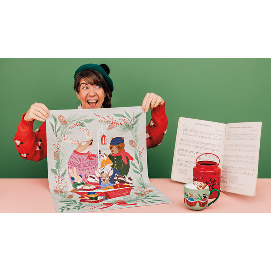 A woman holding up a Woodland Carolers dishtowel by Danica Jubilee next to a matching mug, showing animals singing Christmas Carols.