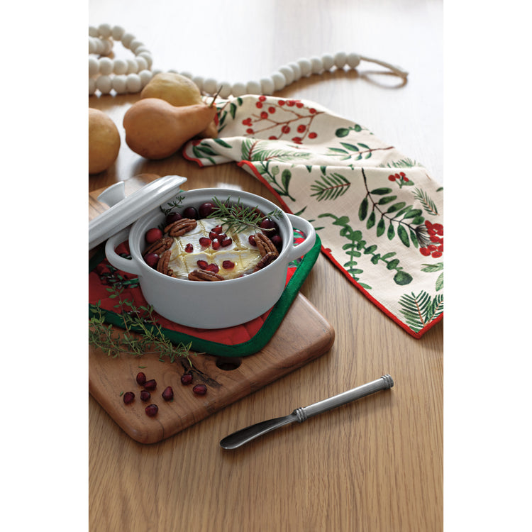 A Now Designs white brie baker with a baked brie inside on an acacia wood cutting board and a Christmas Winterberry printed napkin in the background.