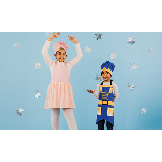 A girl in a Ballerina Kid's Daydream Apron and a boy in a Nutcracker Prince Kid's Daydream Apron by Danica Jubilee with matching chef's hats. The Ballerina apron looks like a pink tutu. They are standing in front of a blue background with snowflakes on it.