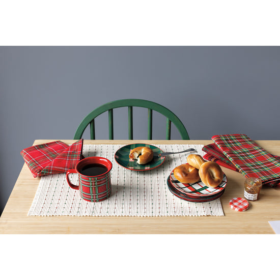 A table with a Now Designs ribbed natural placemat and Christmas plaid napkins, plates, and coffee mug next to a plate of bagels.