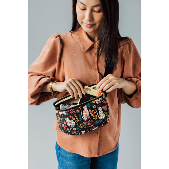 A woman is holding a Cat Bloom hip bag/fanny pack by Danica Studio with cats and flowers on it.