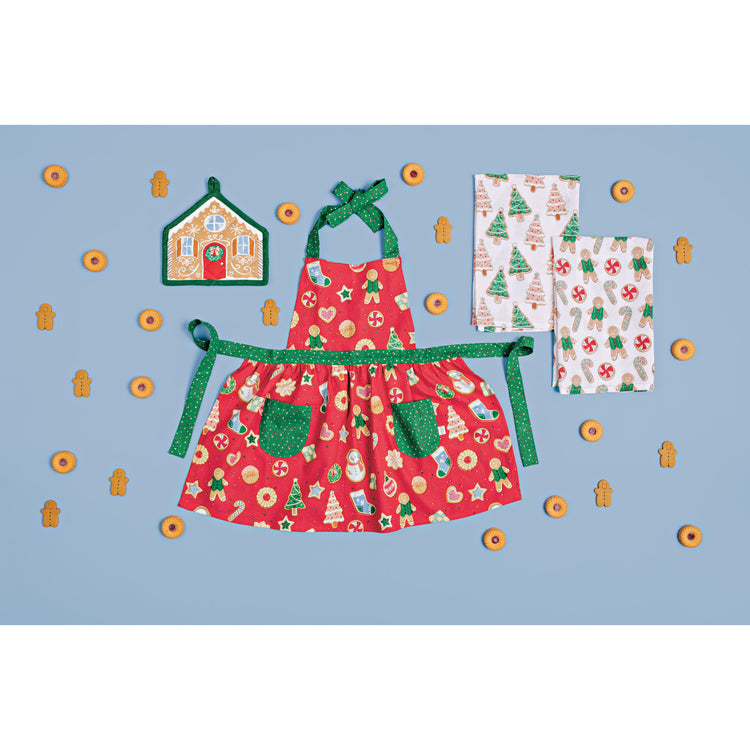 A red printed apron with a gingerbread house shaped potholder and printed dishtowels from the Cookie Exchange Collection by Danica Jubilee, on a blue surface surrounded by Christmas cookies.