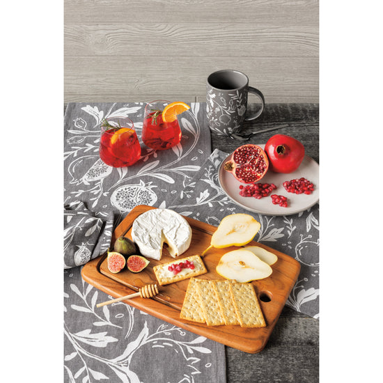 A table with a Now Designs gray and white Pomegranates printed dishtowel and matching mug, and an acacia wood cutting board with crackers, cheese, and fruit on it. There are pomegranates on a plate and red drinks on the table.