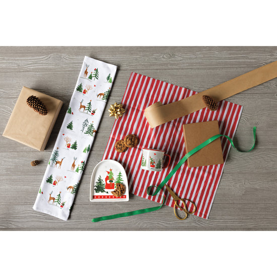 A table with a Now Designs Santa's Reindeer shaped dish and a matching mug and printed dishtowel. There is Christmas gift wrap on the table.