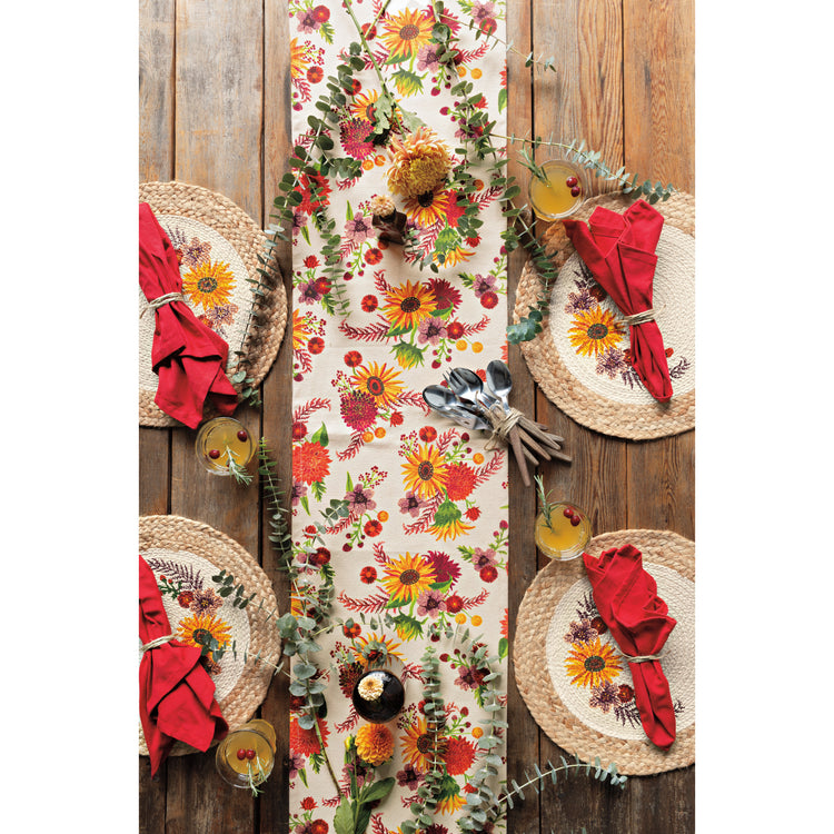 A table setting with Now Designs Sunflower Splendor table linens, including a floral table runner, round braided placemats, and red napkins on a wooden table.