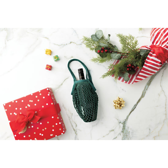 A green Now Designs Le Marché wine tote bag sits on a marble table next to a Christmas gift.