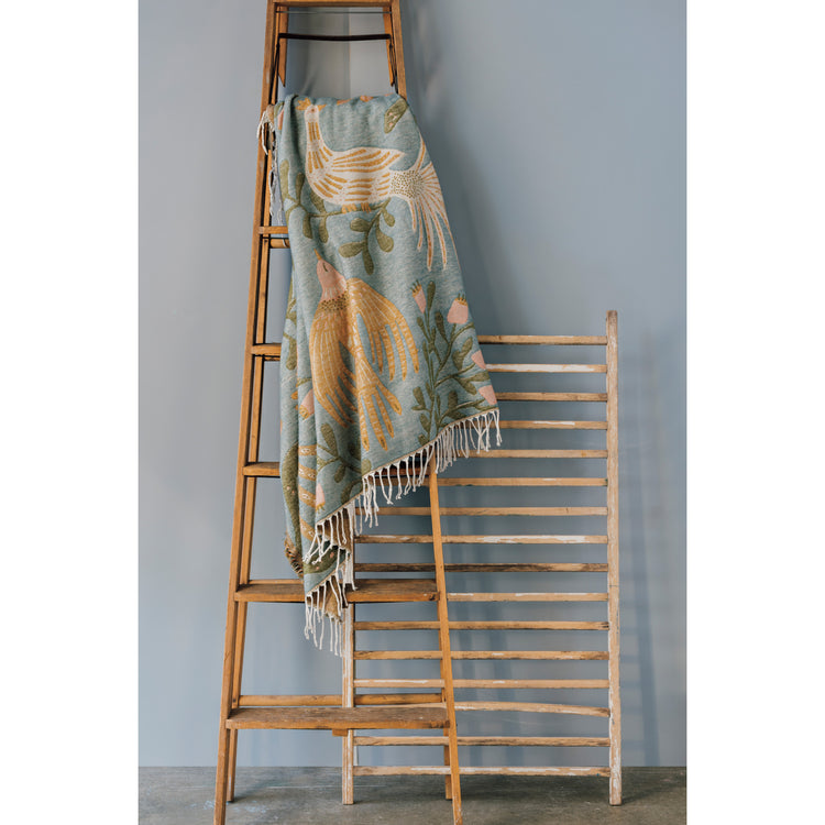 A Danica Studio Plume throw blanket with birds and flowers on it on a wooden ladder next to a blue wall.