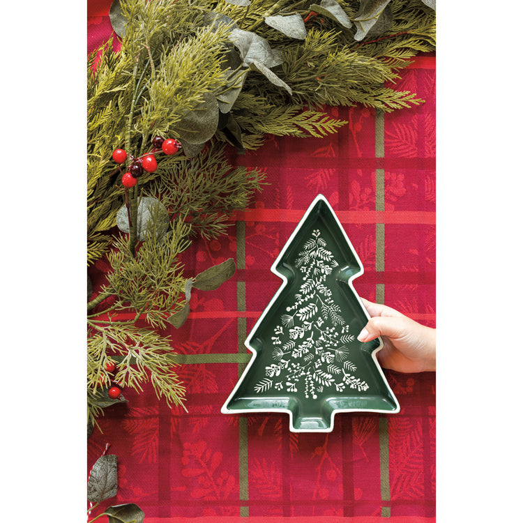 A person holding a Now Designs Winterberry shaped tray in the shape of a Christmas tree, with a plaid Winterberry tablecloth in the background.