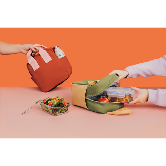 A person holding a Canteloupe green and yellow Danica Jubilee Weekday Lunch Tote with a salad in it and a person holding a Grenadine red and pink Weekday Lunch Tote.