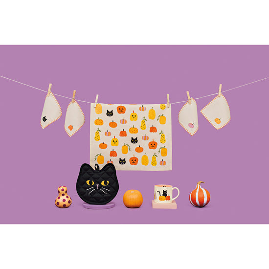 The Hallows' Eve Halloween collection by Danica Jubilee against a purple background, featuring a printed dishtowel with pumpkins on it, cocktail napkins, a black cat shaped potholder, and a mug with a cat and pumpkins on it.