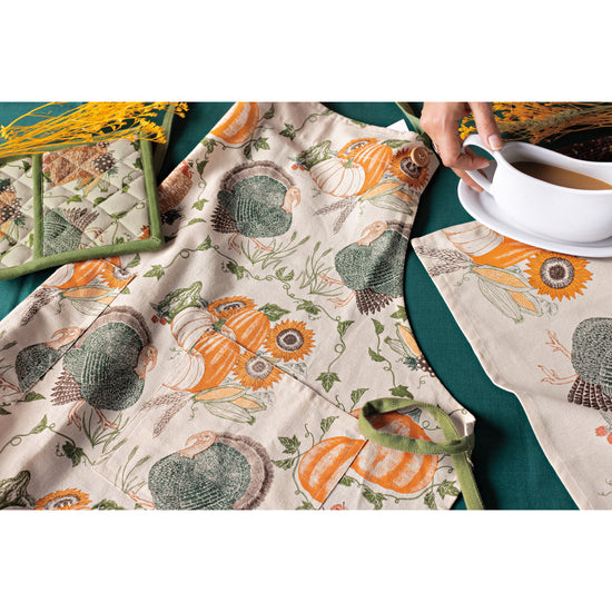 A Now Designs Cornucopia apron with turkeys and pumpkins on it and a white gravy boat on a saucer.