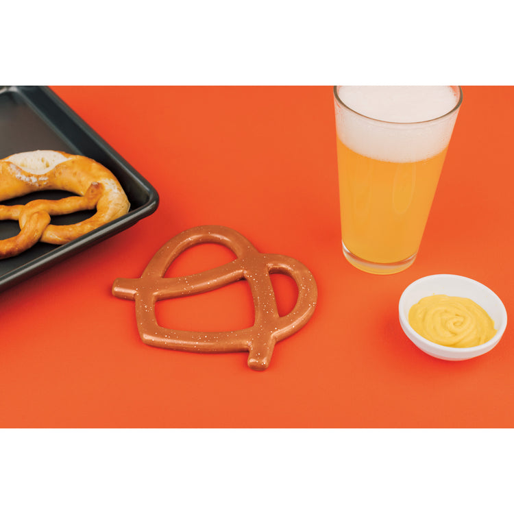 A Danica Jubilee Funny Food pretzel shaped trivet next to a glass of beer.