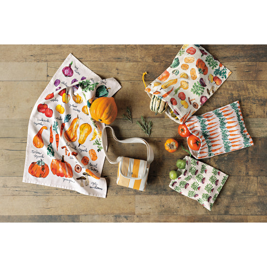 A set of produce bags and a printed dishtowel with vegetables on them from the Now Designs Veggie Stand collection lying on a wooden surface.